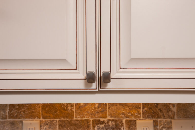 Cream kitchen cabinets with a dark glaze finish and metal knobs above a stone tile backsplash.