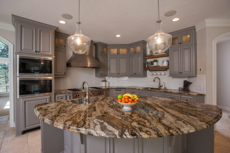 A residential kitchen featuring cabinet painting in warm gray tones, granite island, pendant lights, and stainless applian...