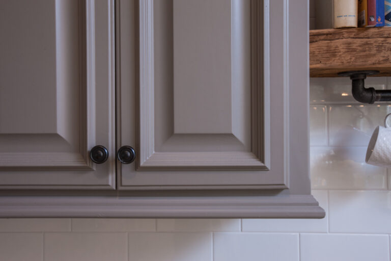 Taupe painted wood kitchen cabinets with decorative molding and black knobs above white subway tile.