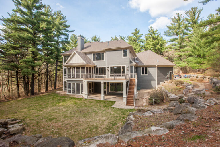 Exterior painting on a large two-story residential home with gray siding, white trim, and wraparound deck surrounded by pi...