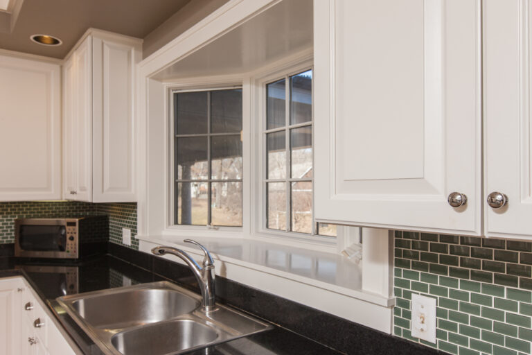 Freshly painted white kitchen cabinets and window trim with a green backsplash and black countertops.