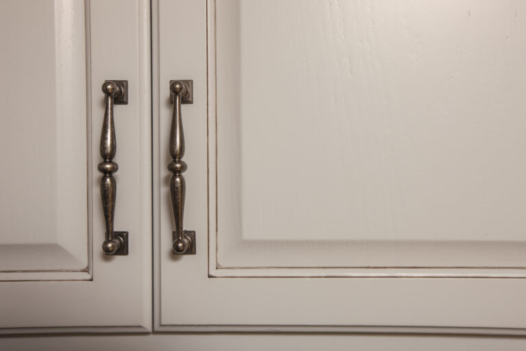 Detail of off-white painted cabinet doors with ornate dark bronze handles and subtle dark glaze.