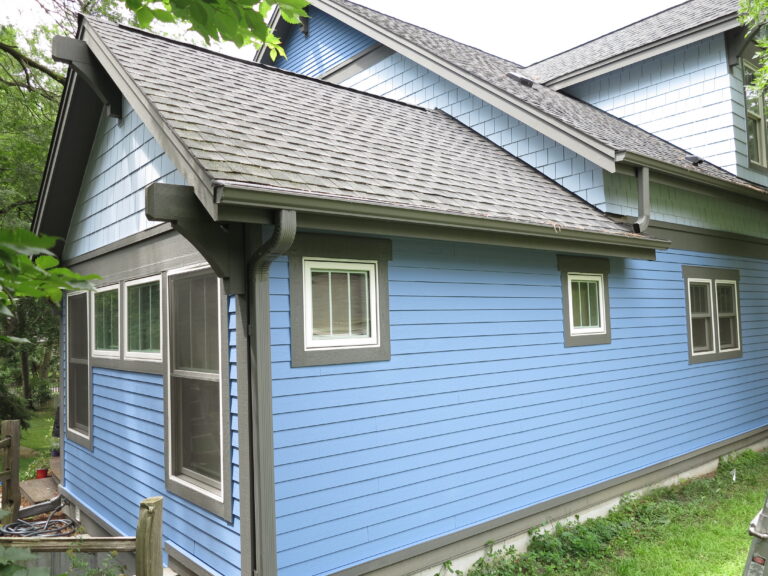 Exterior painting on a residential home with blue lap siding, gray trim, white window frames, and asphalt shingle roof in ...