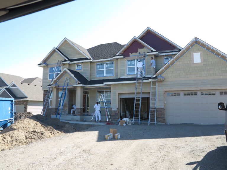Workers on ladders paint the exterior siding and trim of a suburban house at a construction site.