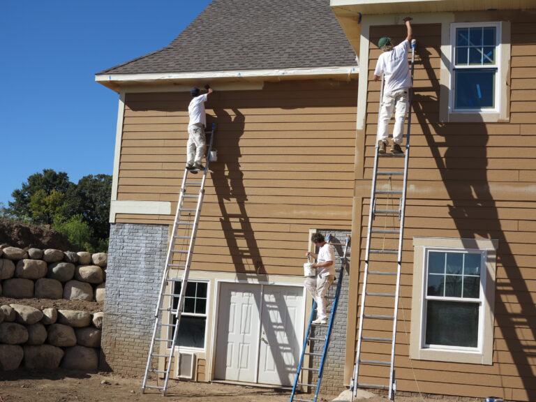 Three painters on tall ladders painting the tan horizontal siding of a house on a sunny day.