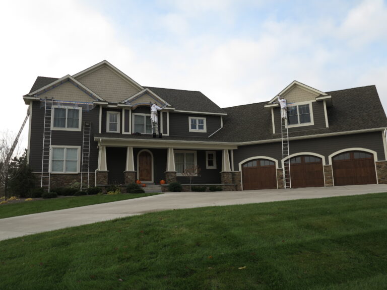 Painters on ladders work on the trim of a large dark gray suburban home with exterior siding.