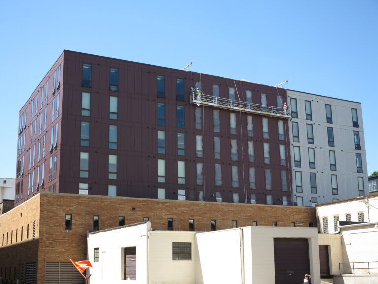 Workers on a suspended scaffold performing exterior painting on a large multi-story commercial building with dark burgundy...