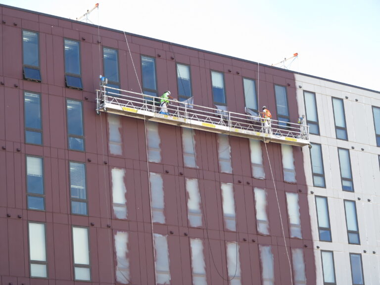 Two workers on a suspended scaffold performing exterior painting on a large multi-story commercial building with dark red ...
