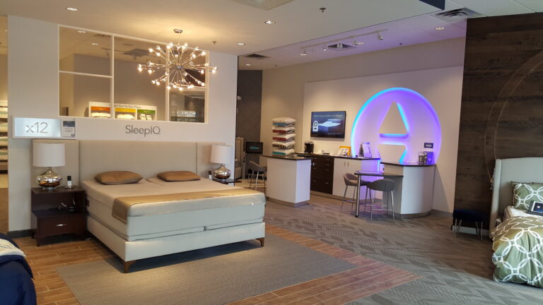 Modern mattress showroom featuring a display bed, branded signage, and a lit logo on a painted wall.