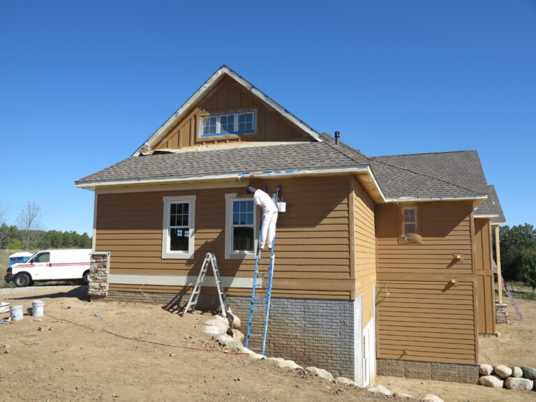 A painter on a ladder doing exterior painting on a new residential home with tan siding and stone foundation trim.