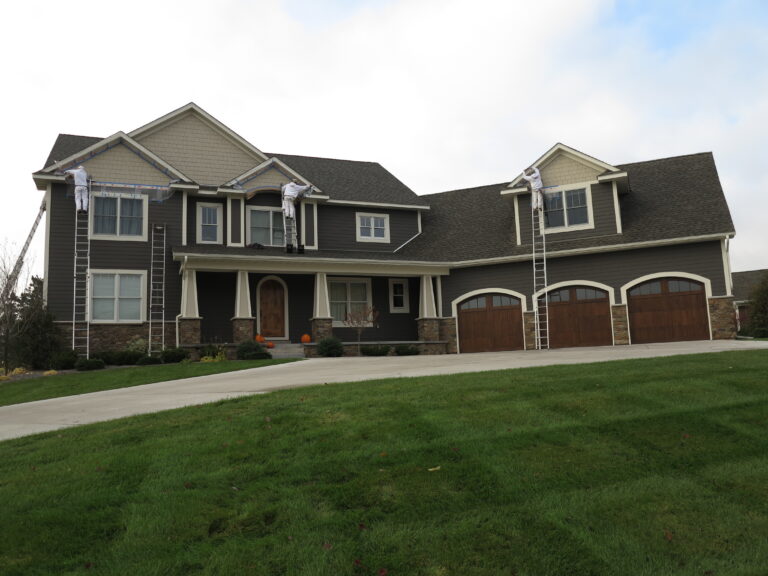 Painters on ladders doing exterior painting on a large two-story residential home with dark gray siding and wood garage do...