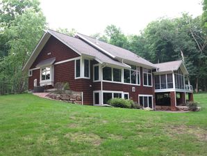 Dark red wood siding and white trim on a hillside house surrounded by lush green trees.