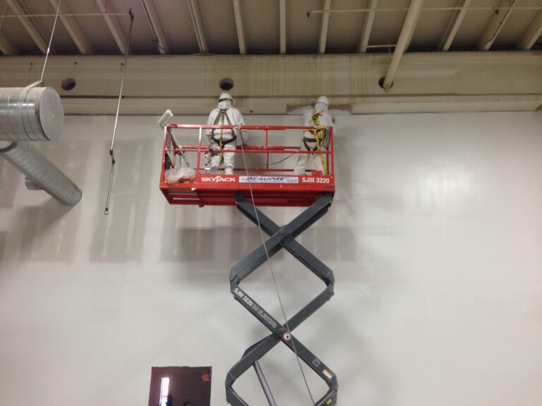 Two painters on a red scissor lift doing commercial interior painting on white walls of a large industrial building.