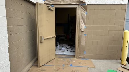 Exterior commercial steel door and block wall being painted tan with protective masking and floor coverings.