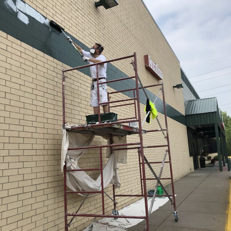 A painter on scaffolding doing exterior painting on a commercial building's dark green and brick facade in Saint Paul.