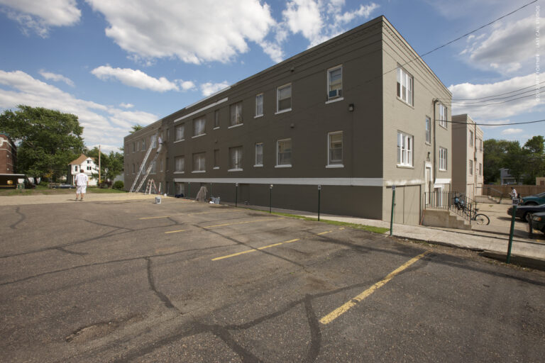 A large three-story brick apartment building being painted gray from a parking lot on a sunny day.